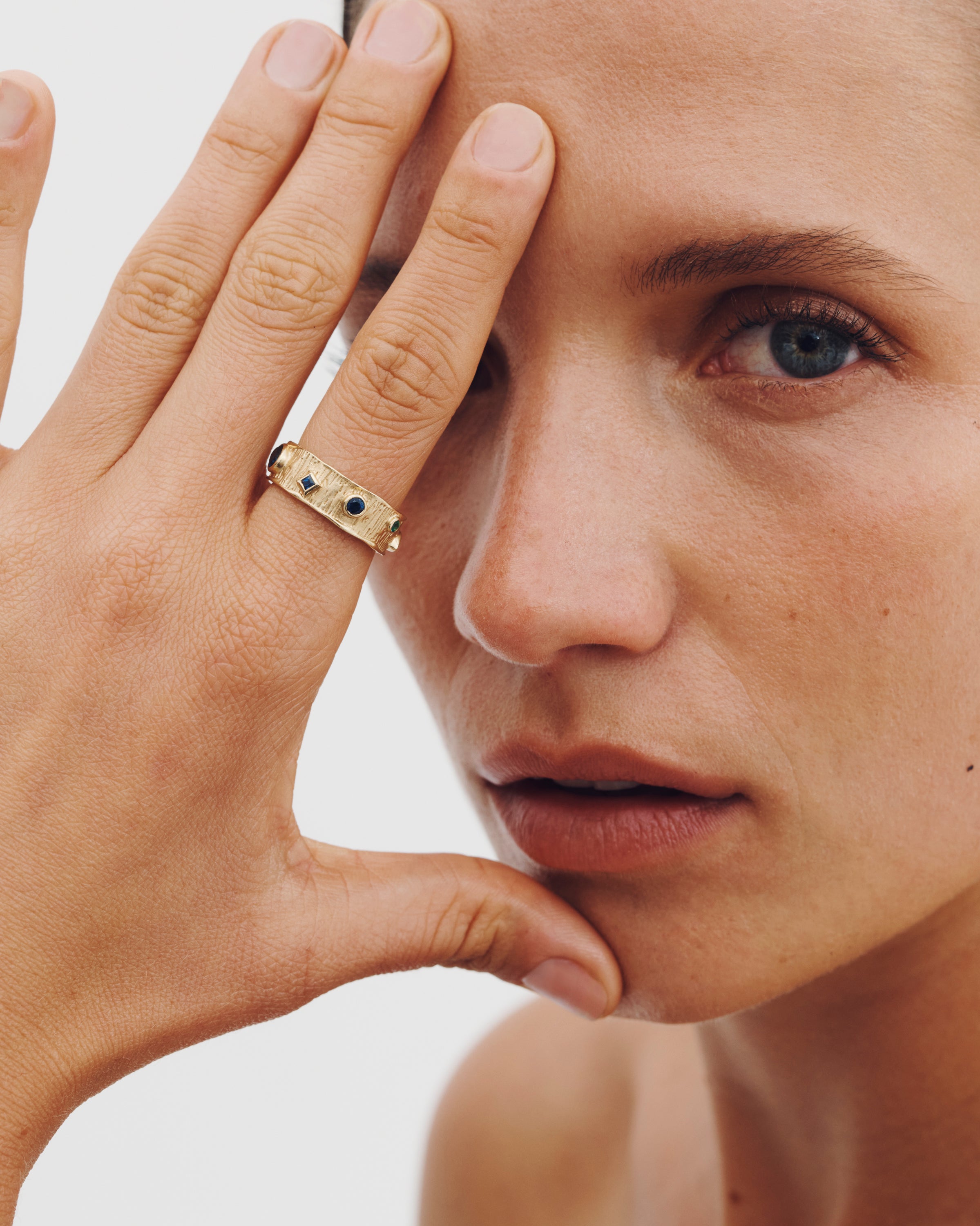 Close-up of a person wearing a gold ring with blue stones, holding their hand near their face.