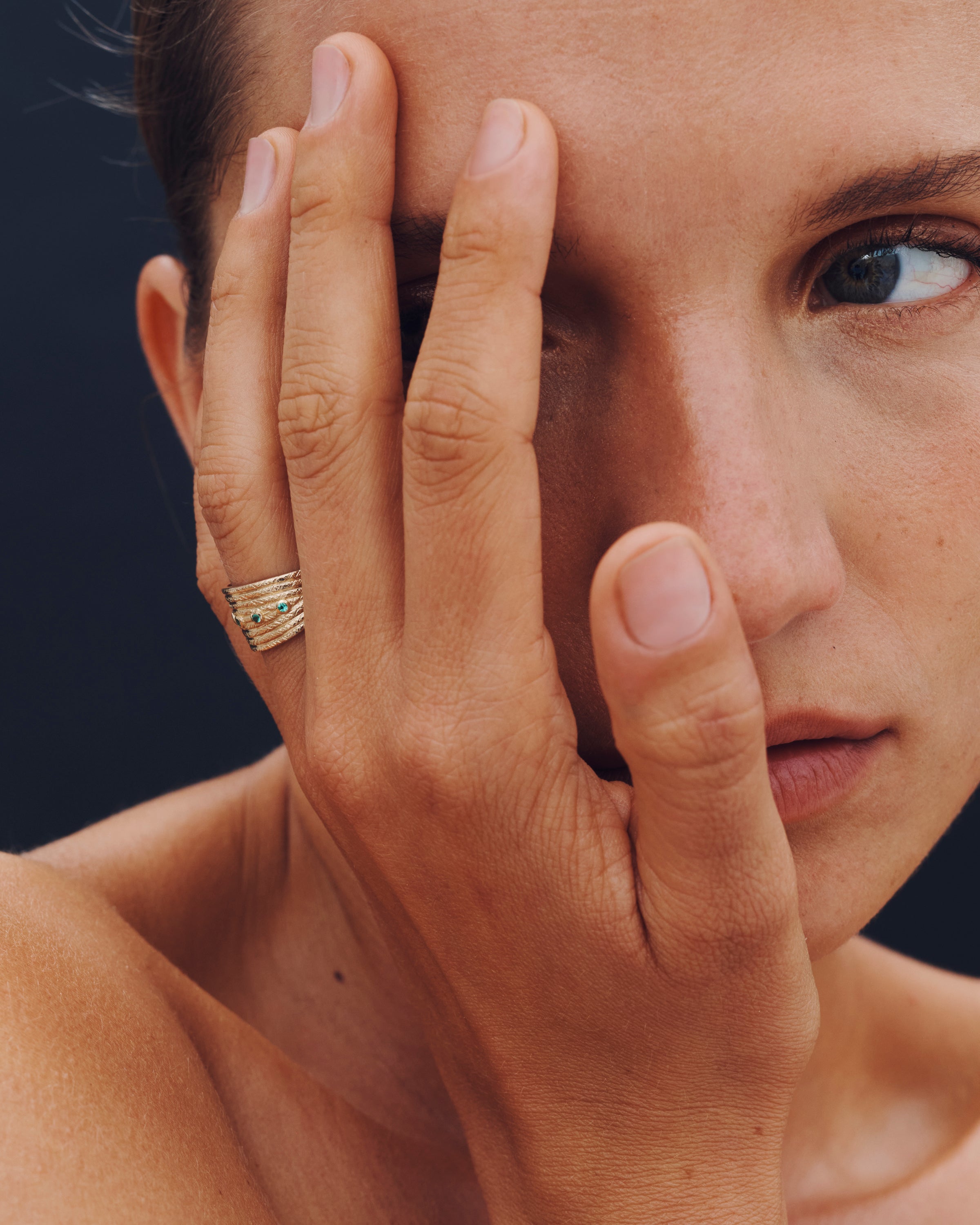Woman with a gold ring on her finger holding her hand up to her face against a dark background