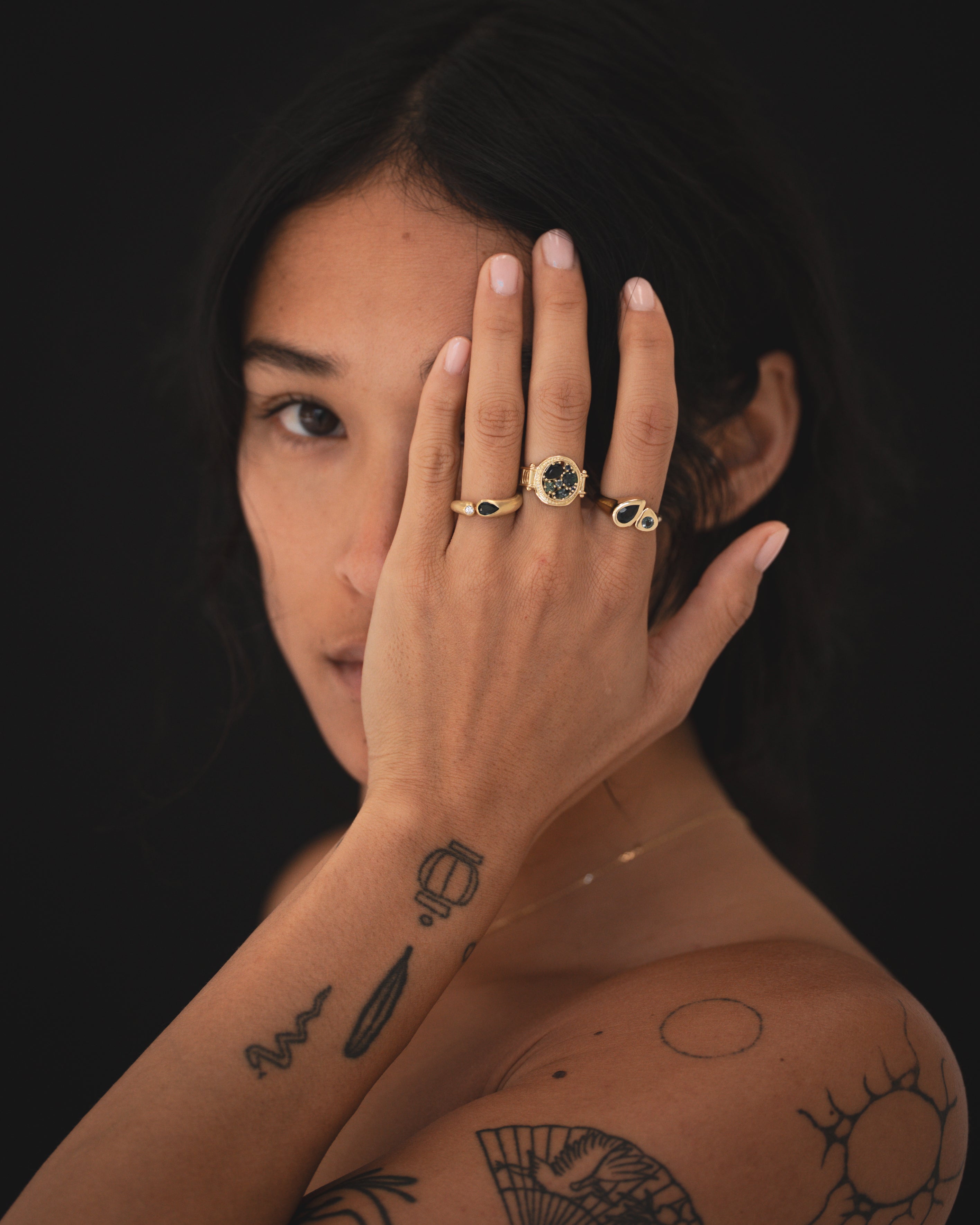 Woman with tattoos wearing two gold rings on a dark background
